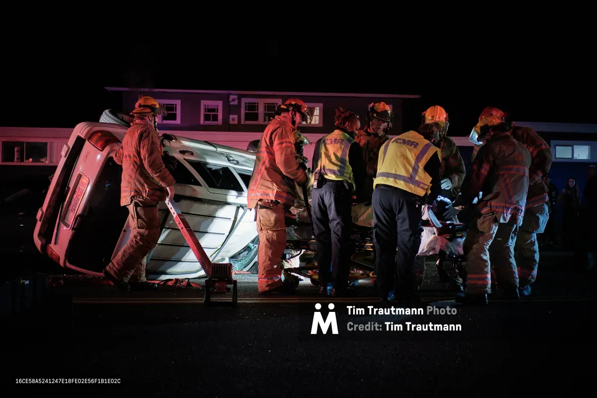 Portland firefighters and paramedics work urgently around an overturned silver SUV on Northeast Killingsworth Street in the Humboldt neighborhood. The scene is bathed in dramatic red and blue emergency lighting against the stark black night, creating an intense atmosphere of coordinated rescue efforts. Multiple first responders in high-visibility gear and protective helmets cluster around the damaged vehicle, their movements deliberate and focused as they tend to injured passengers. The residential backdrop of modest homes provides a sobering contrast to the chaos of twisted metal and emergency response.