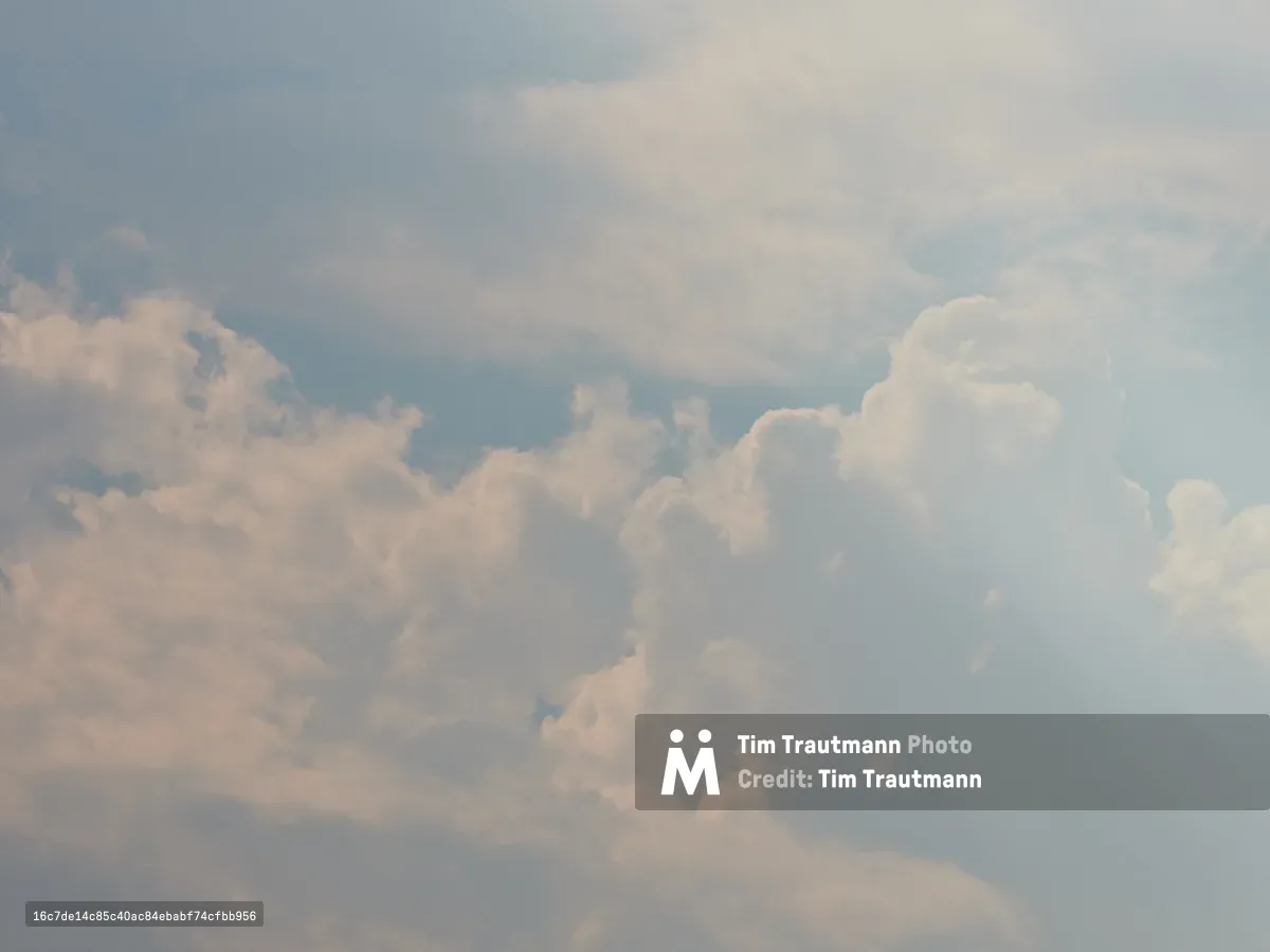A dramatic sky filled with billowing white and gray cumulus clouds against a soft blue backdrop, captured over Portland, Oregon. The clouds create layers of depth and texture across the entire frame, suggesting changing weather conditions in the Pacific Northwest.