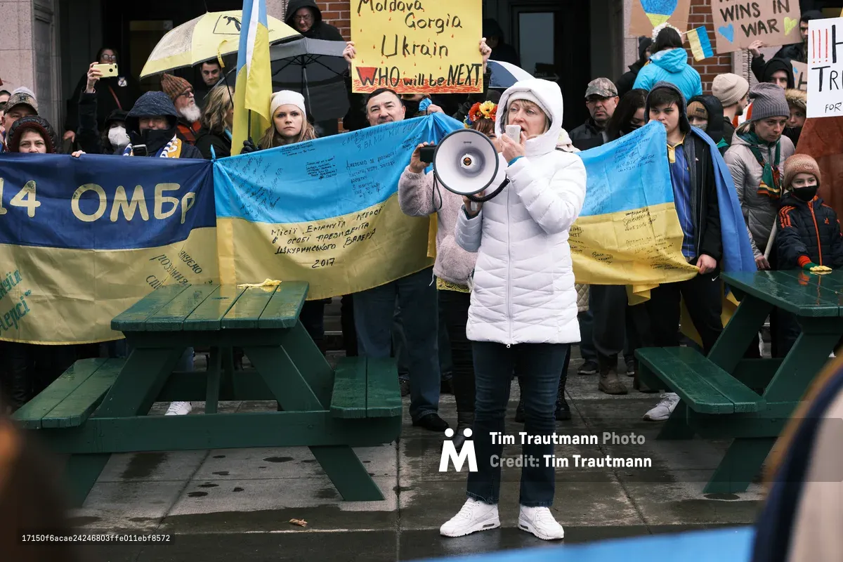A determined protester in a white puffy jacket amplifies her message through a megaphone, standing before a weathered Ukrainian flag inscribed with signatures and messages of support. The scene unfolds on the rain-dampened concrete plaza outside Portland's Revolution Hall, where dozens of bundled demonstrators have gathered in the gray winter light. Handmade signs warning "WHO will be NEXT" pierce through the crowd, creating a prophetic tableau of solidarity captured just days before Russia's invasion began.