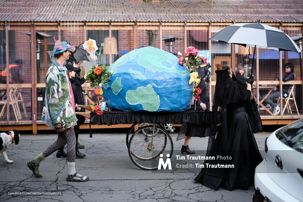 A group of people in mourning attire participate in an environmental protest or performance art piece, wheeling a large painted Earth globe on a funeral cart through the streets of Portland's Central Eastside district. The participants wear black clothing and carry flowers, creating a somber funeral procession for the planet.