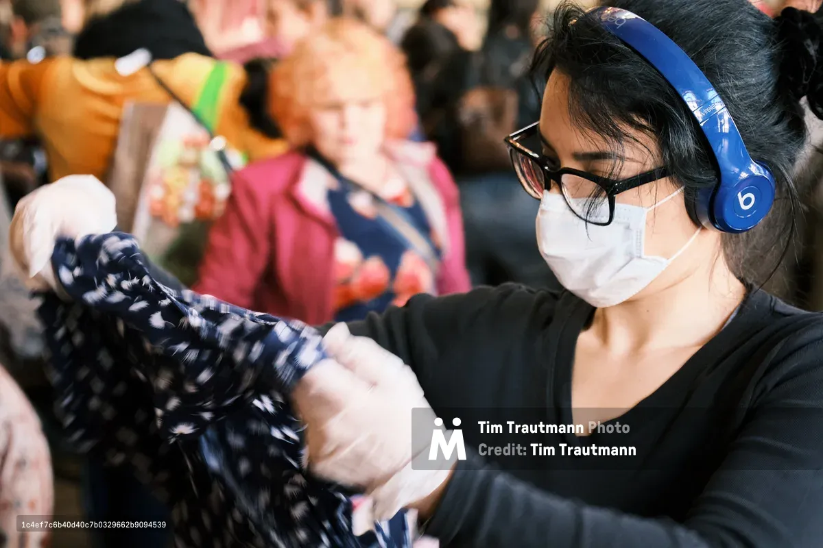 A shopper wearing a white face mask, black-rimmed glasses, and blue Beats wireless headphones examines a patterned garment at a crowded swap meet inside the Crystal Ballroom in Portland, Oregon. She wears a black long-sleeve top and is focused on inspecting the clothing item. A busy crowd of other shoppers is visible in the blurred background.