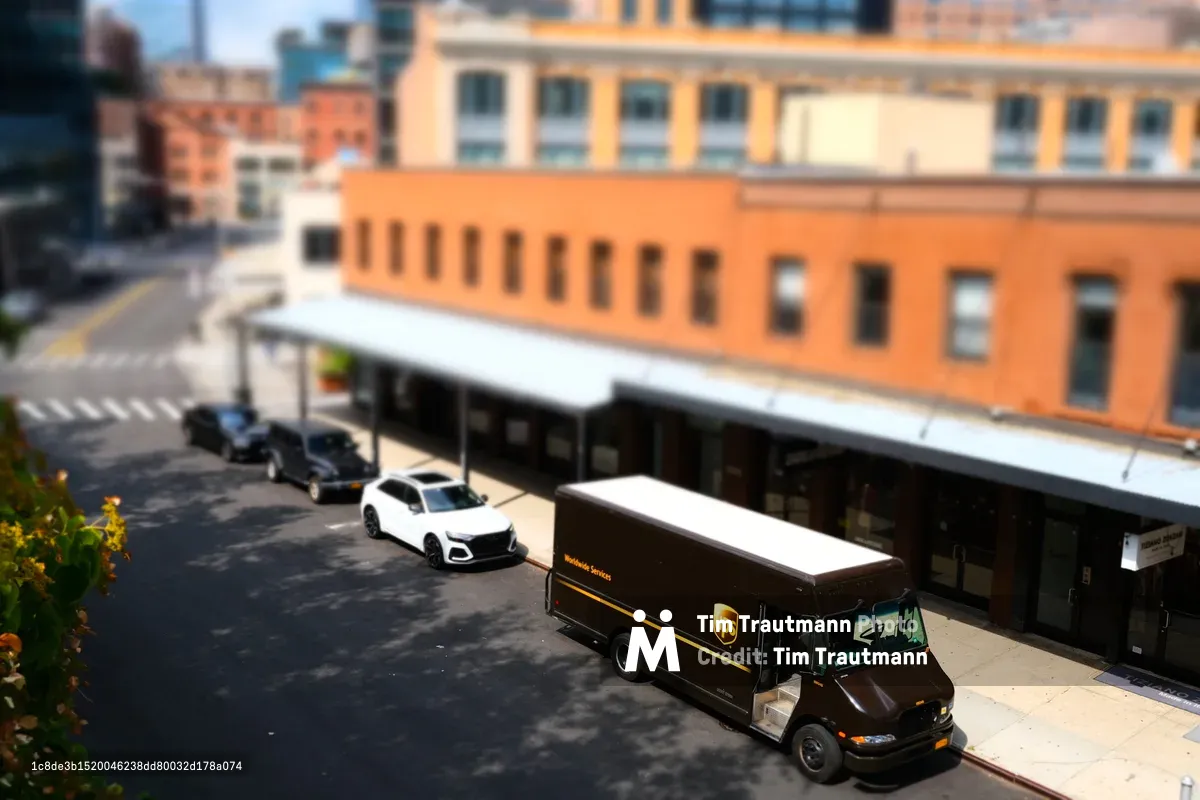 A chocolate-brown UPS delivery truck navigates the sun-dappled street below Manhattan's elevated High Line park, captured with dreamy tilt-shift photography that transforms the Meatpacking District scene into a toy-like diorama. The selective focus creates an enchanting miniature effect, with warm terracotta and cream-colored industrial buildings providing a textured backdrop. Shadows dance across the asphalt while a white sedan waits patiently, the entire urban tableau bathed in golden afternoon light that accentuates the architectural details of this transformed neighborhood.