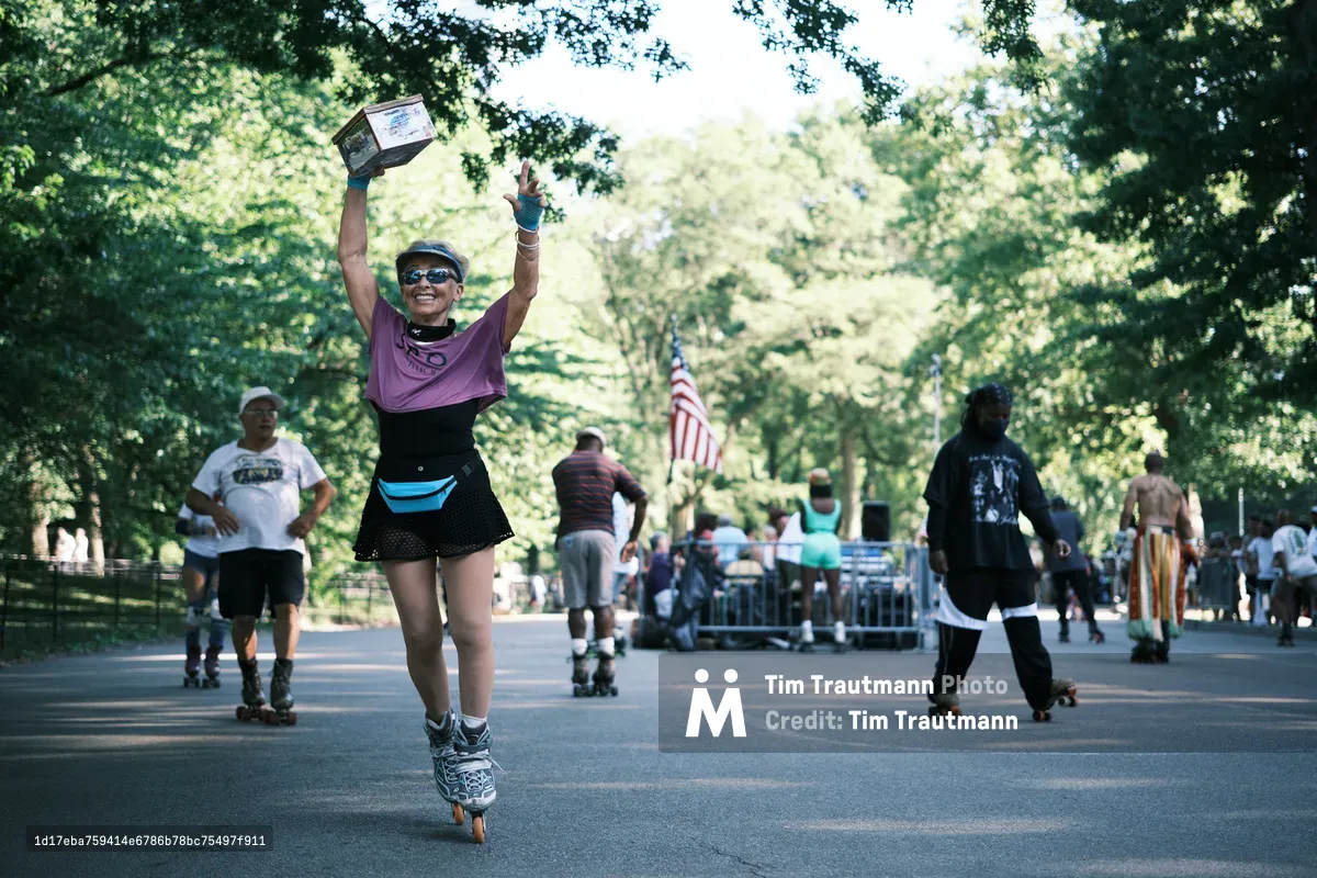 A jubilant roller skater in purple and black athletic wear celebrates triumphantly on Central Park's tree-lined pathways, both arms raised high while clutching what appears to be an award or trophy. Dappled sunlight filters through the dense summer canopy, creating a luminous backdrop as other skaters glide leisurely in the distance. The American flag stands proudly among the verdant trees, while the smooth asphalt stretches invitingly ahead, capturing the pure joy of recreational skating in Manhattan's beloved green heart.