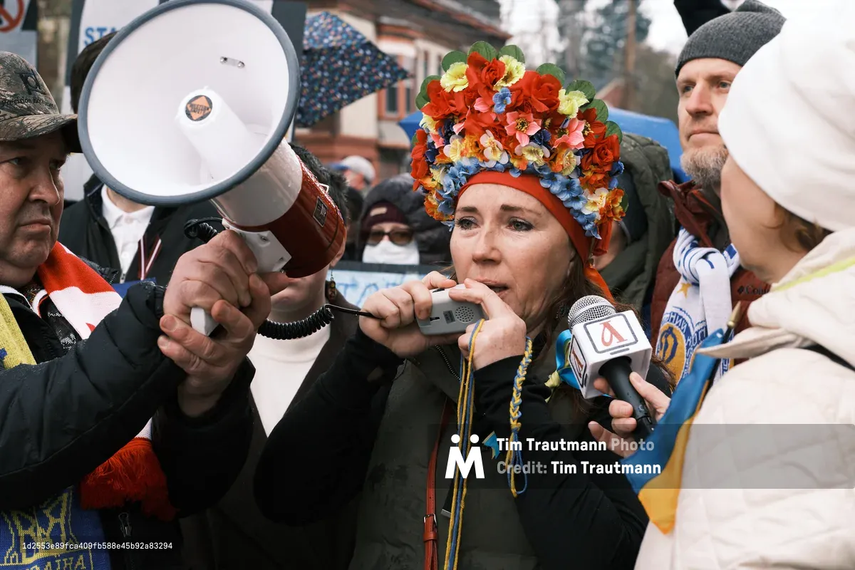 A woman crowned with an elaborate headdress of vibrant artificial flowers—red roses mingling with blue and yellow blooms—speaks into multiple microphones thrust toward her by surrounding media and protesters. Her weathered face reflects determination as she addresses the crowd outside Portland's Revolution Hall, wrapped in dark winter clothing against the gray February chill. The scene pulses with urgent energy as megaphones and recording devices create a constellation of amplification around her impassioned words, while Ukrainian flag colors echo symbolically in her floral crown.