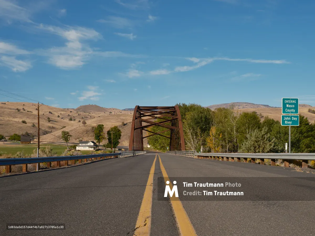 A weathered steel truss bridge spans the John Day River along the Shaniko-Fossil Highway, marking the boundary between Wheeler and Wasco counties in Oregon's high desert. Golden afternoon light bathes the rolling, sage-dotted hills while wispy cirrus clouds stretch across an expansive blue sky. The perspective from the center of the asphalt road, with its worn yellow dividing lines, draws the eye through the russet bridge structure toward distant homesteads nestled in the fold of ancient hills.