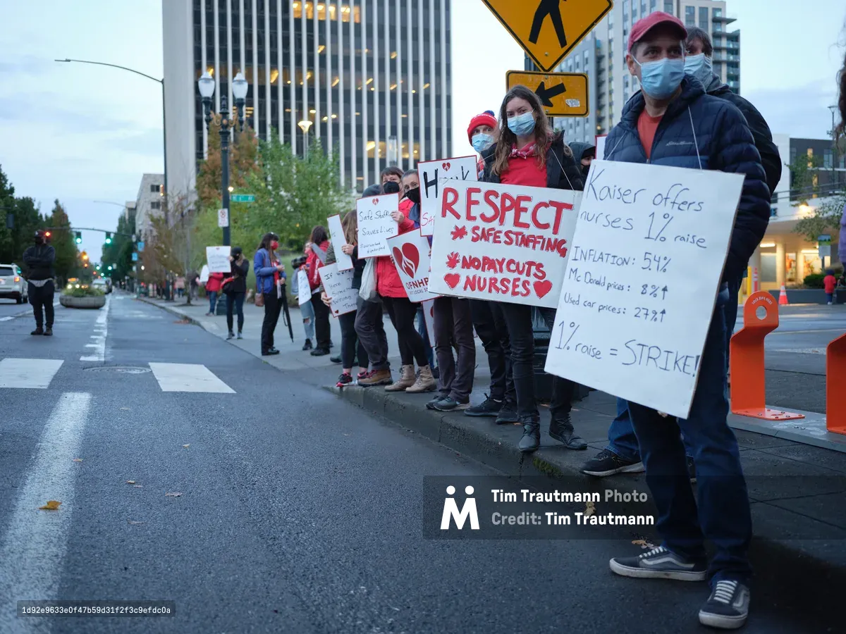 Healthcare workers line the rain-slicked streets of Portland's Lloyd District, their handwritten protest signs catching the amber glow of evening streetlights against towering medical office buildings. Masked demonstrators in scrubs and casual wear hold placards demanding respect, safe staffing levels, and fair compensation, their determined faces reflecting the gravity of their cause. The wet pavement mirrors the urban canyon of glass and steel that houses Kaiser Permanente's regional headquarters, creating a stark juxtaposition between corporate power and grassroots advocacy. The photographer captures this moment of collective action with intimate street-level perspective, emphasizing the human scale of the protest against the institutional backdrop.