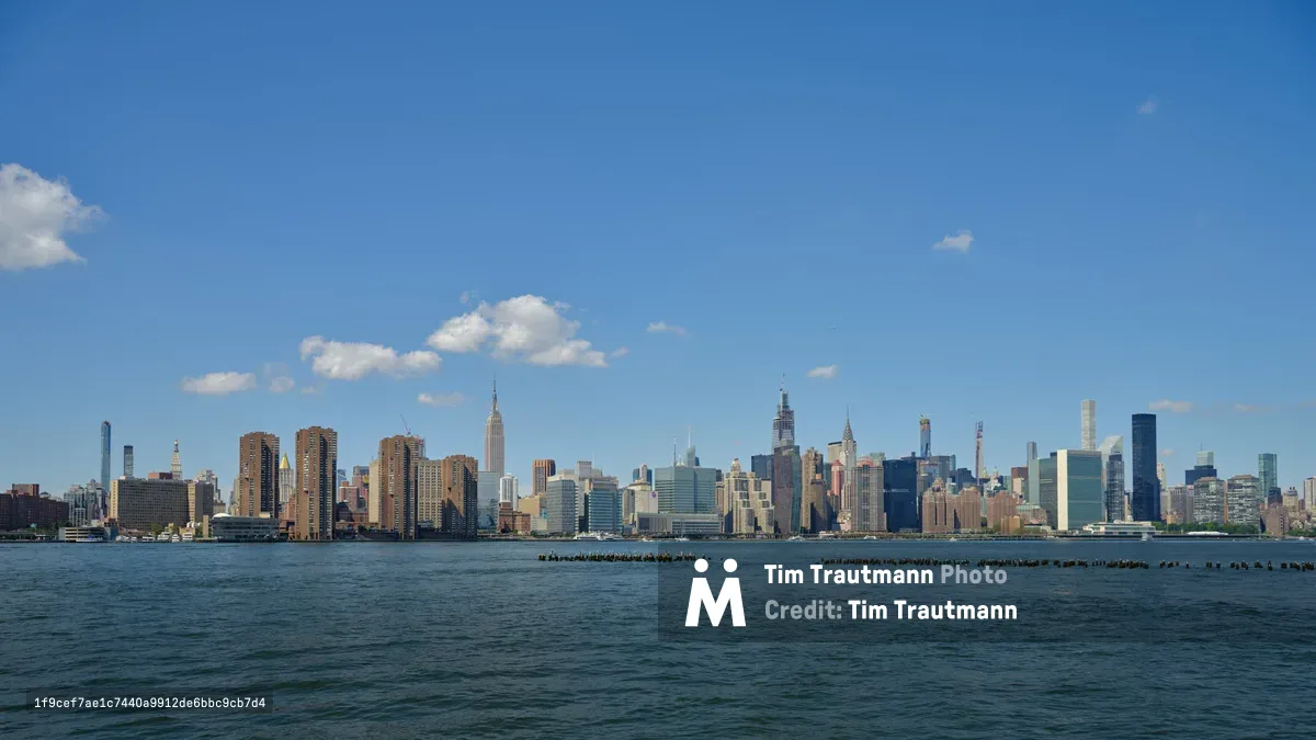 The iconic Manhattan skyline rises majestically across the East River, viewed from the industrial waterfront of Greenpoint, Brooklyn. Wispy white clouds drift across an azure afternoon sky, creating a serene backdrop for the dense cluster of skyscrapers including the distinctive Empire State Building. The foreground features weathered wooden pier pilings emerging from the dark river waters, adding rustic texture to this classic New York vista. The composition captures the dramatic contrast between Brooklyn's quieter shores and Manhattan's towering urban density.