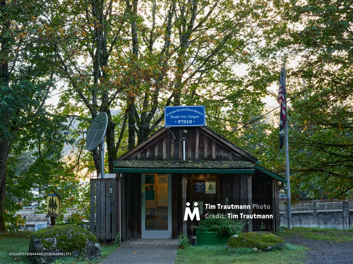 A weathered wooden post office building sits nestled beneath towering deciduous trees in the late afternoon light of Bridal Veil, Oregon. The rustic structure features vertical board siding darkened by time and moss, with a distinctive blue USPS sign marking it as zip code 97010. An American flag stands sentinel beside the humble building, while dappled sunlight filters through the canopy above, creating a nostalgic atmosphere that speaks to America's vanishing small-town postal heritage.