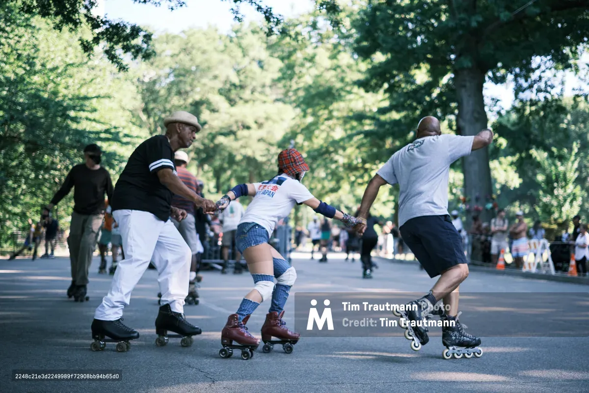 Three generations converge in a spirited tug-of-war beneath Central Park's dappled canopy, their determination etched in strained postures and interlocked grips. An elderly man in a beret anchors one end while a young participant in roller skates and tartan provides the middle link, connected to a man in inline skates who leans into the effort. Filtered sunlight streams through the verdant foliage, casting dancing shadows across the park's asphalt pathway where weekend recreation unfolds.