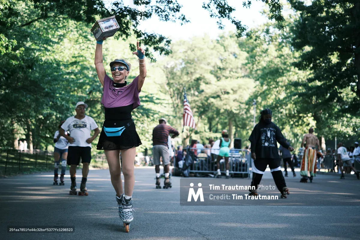 A triumphant roller skater in purple athletic wear raises both arms in celebration while gliding down Central Park's tree-lined pathway, a trophy box held high above her head. The scene captures pure joy as filtered sunlight creates dancing patterns through the dense summer canopy, while fellow skaters and park visitors populate the background beneath an American flag. The composition draws the eye to her moment of victory against the park's iconic verdant tunnel, creating an atmosphere of community celebration and athletic achievement.