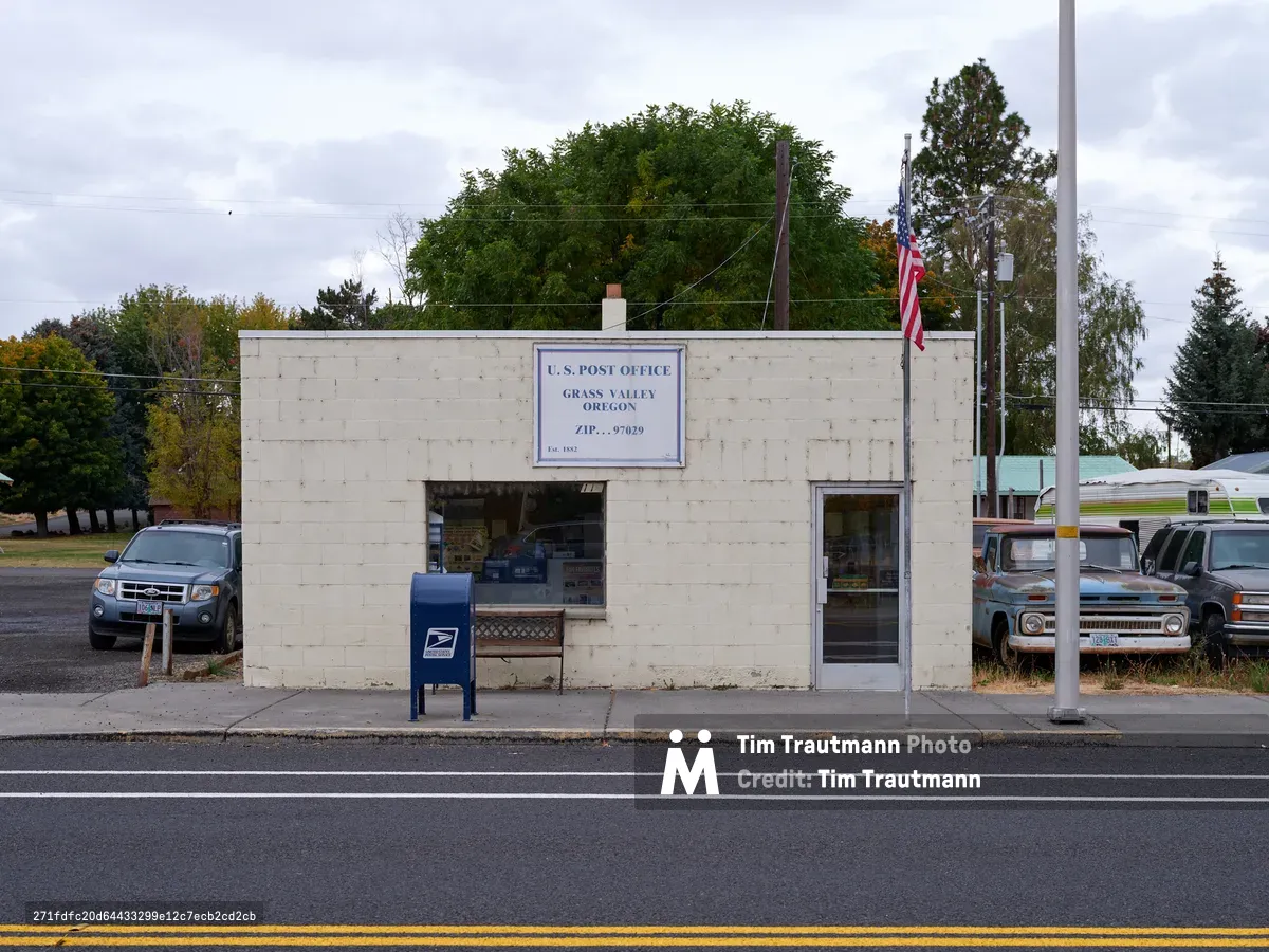 A modest concrete block U.S. Post Office stands sentinel along Sherman Highway in rural Grass Valley, Oregon, its weathered white facade bearing the ZIP code 97029 beneath overcast autumn skies. The utilitarian building, flanked by aging pickup trucks and a solitary blue mailbox, embodies the enduring spirit of small-town America where community connections persist through simple civic institutions. Power lines stretch overhead while mature trees provide a verdant backdrop to this quintessentially rural Pacific Northwest scene.