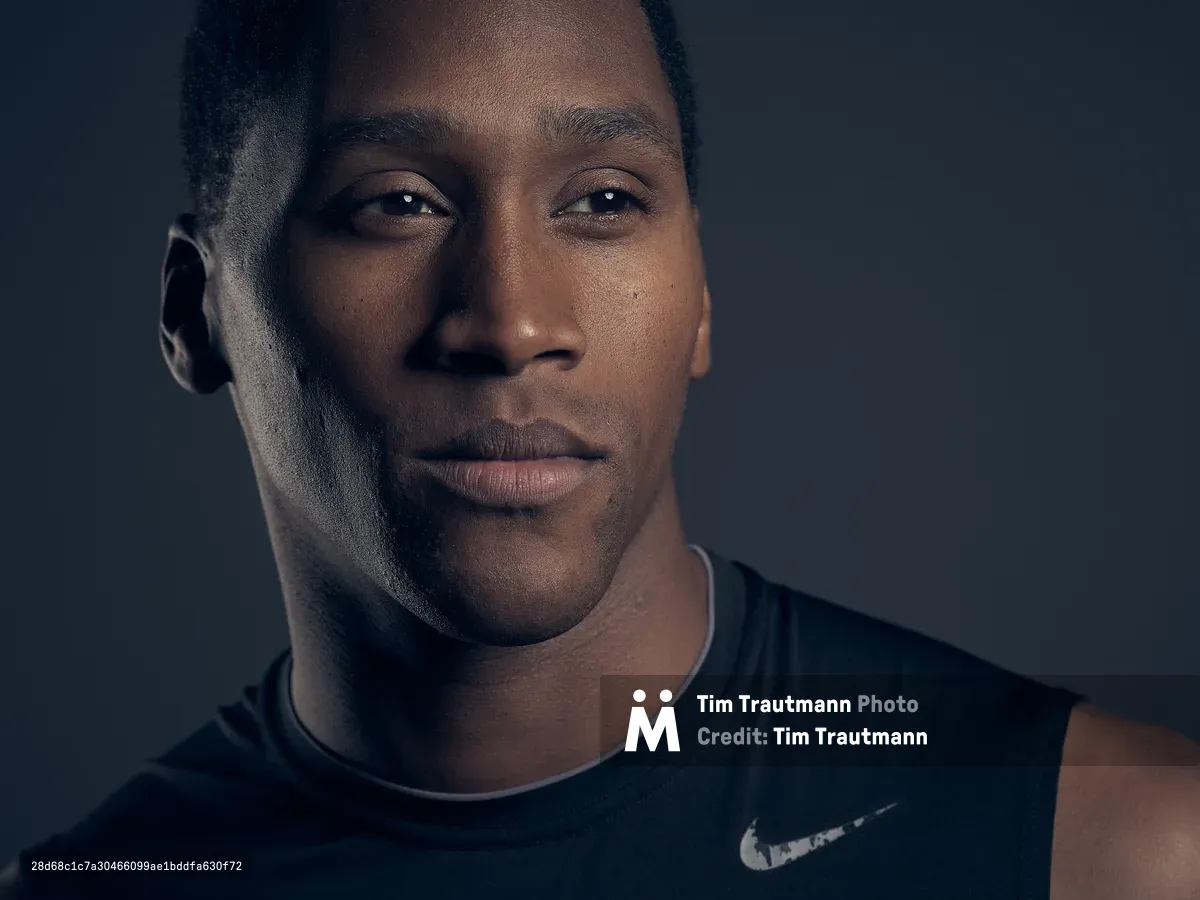 A close-up studio portrait of a young man wearing a black Nike sleeveless shirt, lit dramatically against a dark grey background. He gazes slightly off-camera with a calm, composed expression.