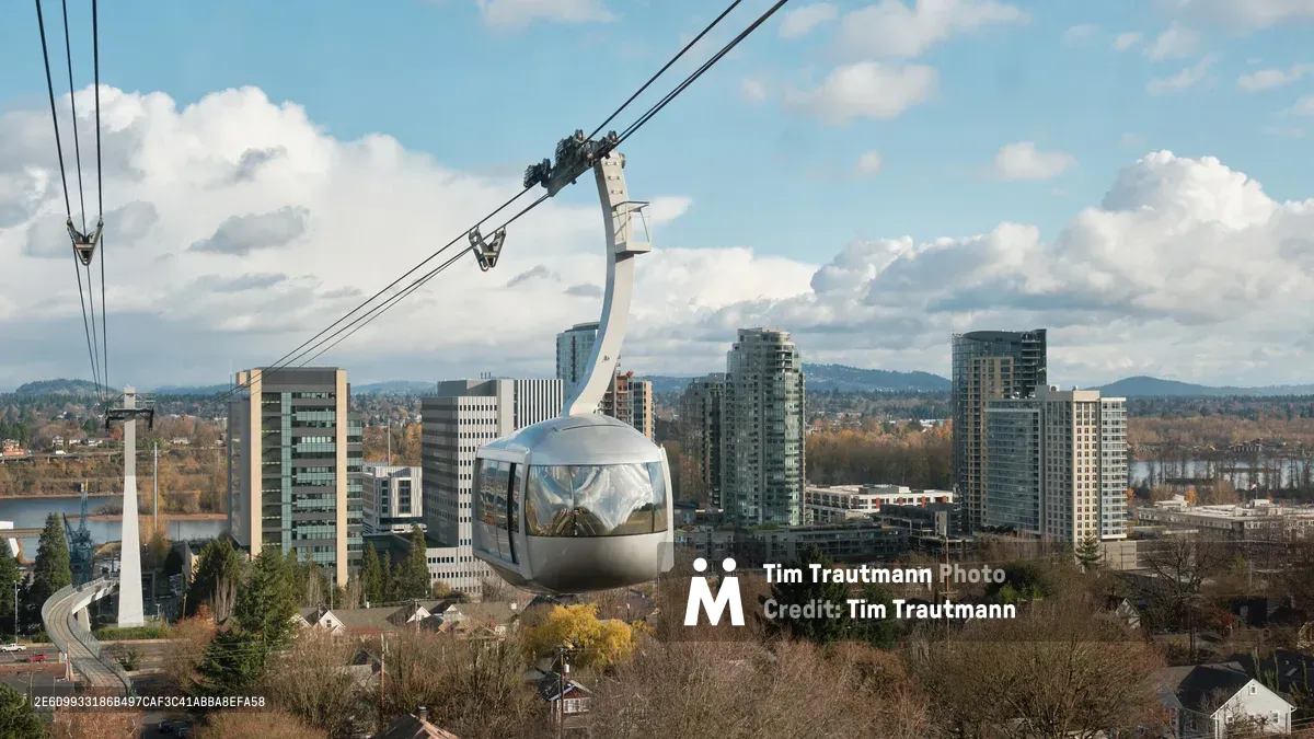 A sleek silver gondola car of the Portland Aerial Tram floats gracefully through autumn air, its reflective surface catching the soft overcast light above the South Waterfront district. The panoramic view reveals Portland's modern high-rise towers nestled between the Willamette River and rolling hills, while bare-branched trees in the foreground signal late fall's arrival. The composition captures both the engineering marvel of the tram system and the urban landscape it serves, connecting OHSU's Marquam Hill campus to the bustling waterfront below.
