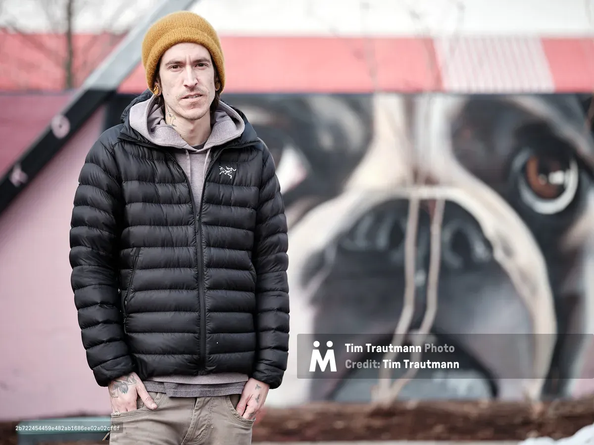 A portrait of muralist Kyle Danley, also known as Wetiko, standing in front of one of his large-scale murals depicting a close-up of a French Bulldog's face on a pink building. He wears a black Arc'teryx puffer jacket, a grey hoodie, khaki jeans, and a mustard yellow beanie. Neck and hand tattoos are visible. He looks directly into the camera with a calm, serious expression, hands in his pockets.