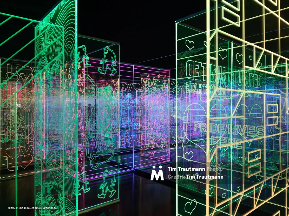 A vibrant interactive art installation featuring glowing neon outlines in green, purple, and blue colors creating geometric patterns, text, and heart shapes against a dark background. The illuminated display creates an immersive digital art experience with layered transparent panels and wireframe designs.