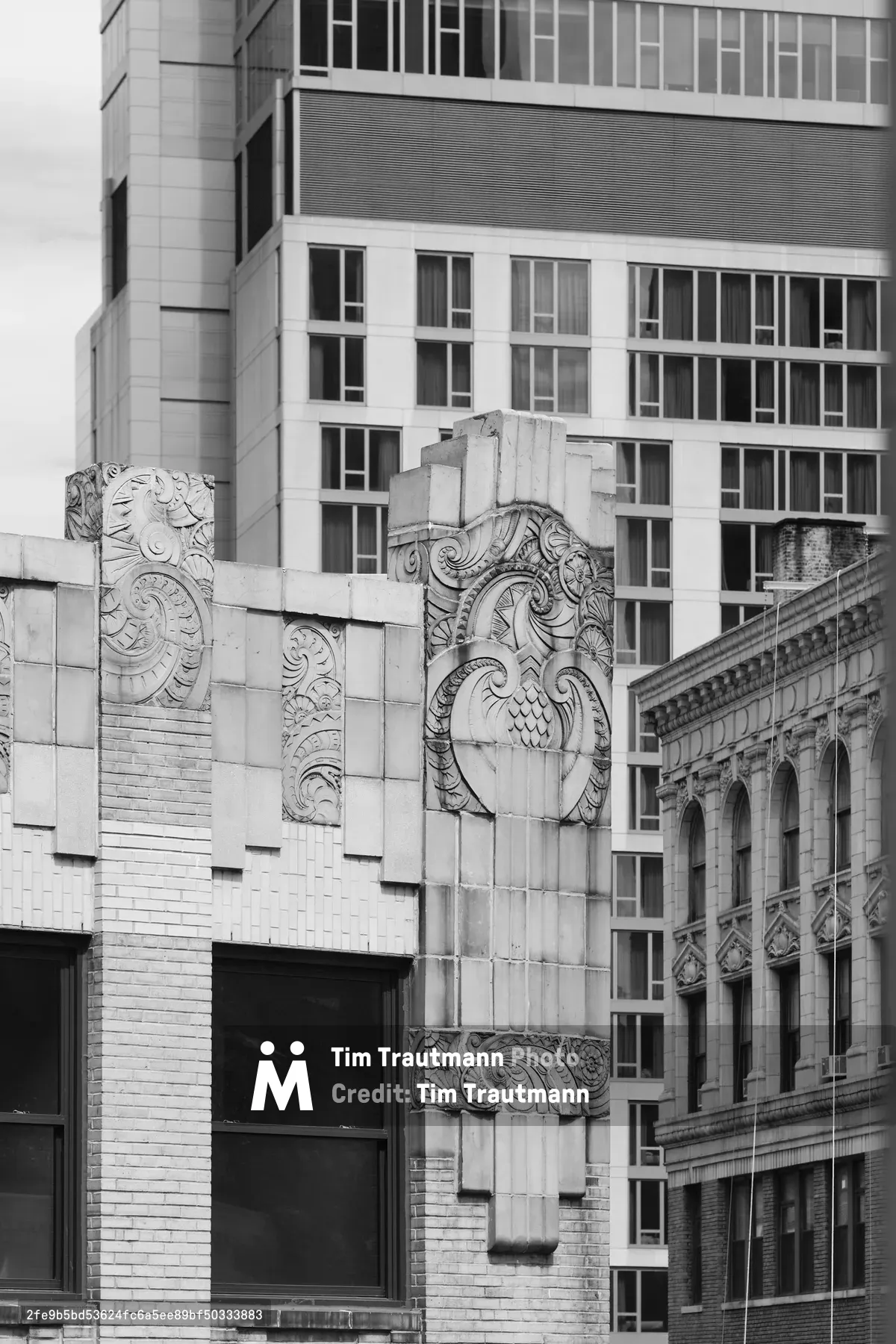 Elaborate carved stone ornaments crown the corner of a historic building in Manhattan's NoMad district, their intricate Art Deco flourishes creating dramatic shadows against weathered limestone. The monochromatic composition captures the interplay between ornate early 20th-century craftsmanship and the stark modernist glass tower rising behind, while classical arched windows peek from the right frame. Soft, diffused light emphasizes the sculptural depth of the decorative elements, revealing every carved detail in the building's ornamental crown.