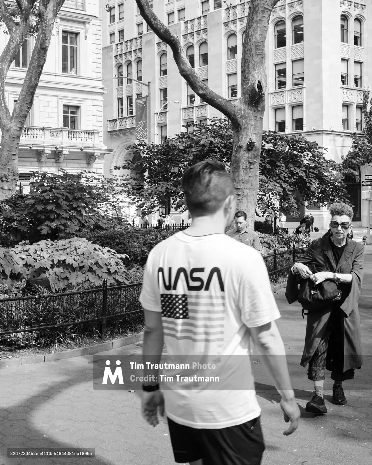 A figure in a NASA t-shirt with American flag graphic walks through Madison Square Park in the Flatiron District, their motion creating a gentle blur against the static urban tableau. An elderly man in dark coat and sunglasses sits contemplatively on a bench, framed by the ornate Beaux-Arts and Renaissance Revival architecture that defines this historic Manhattan neighborhood. The dappled shadows of mature plane trees create a natural canopy over the scene, while the classical facades of the surrounding buildings speak to New York's gilded age grandeur.