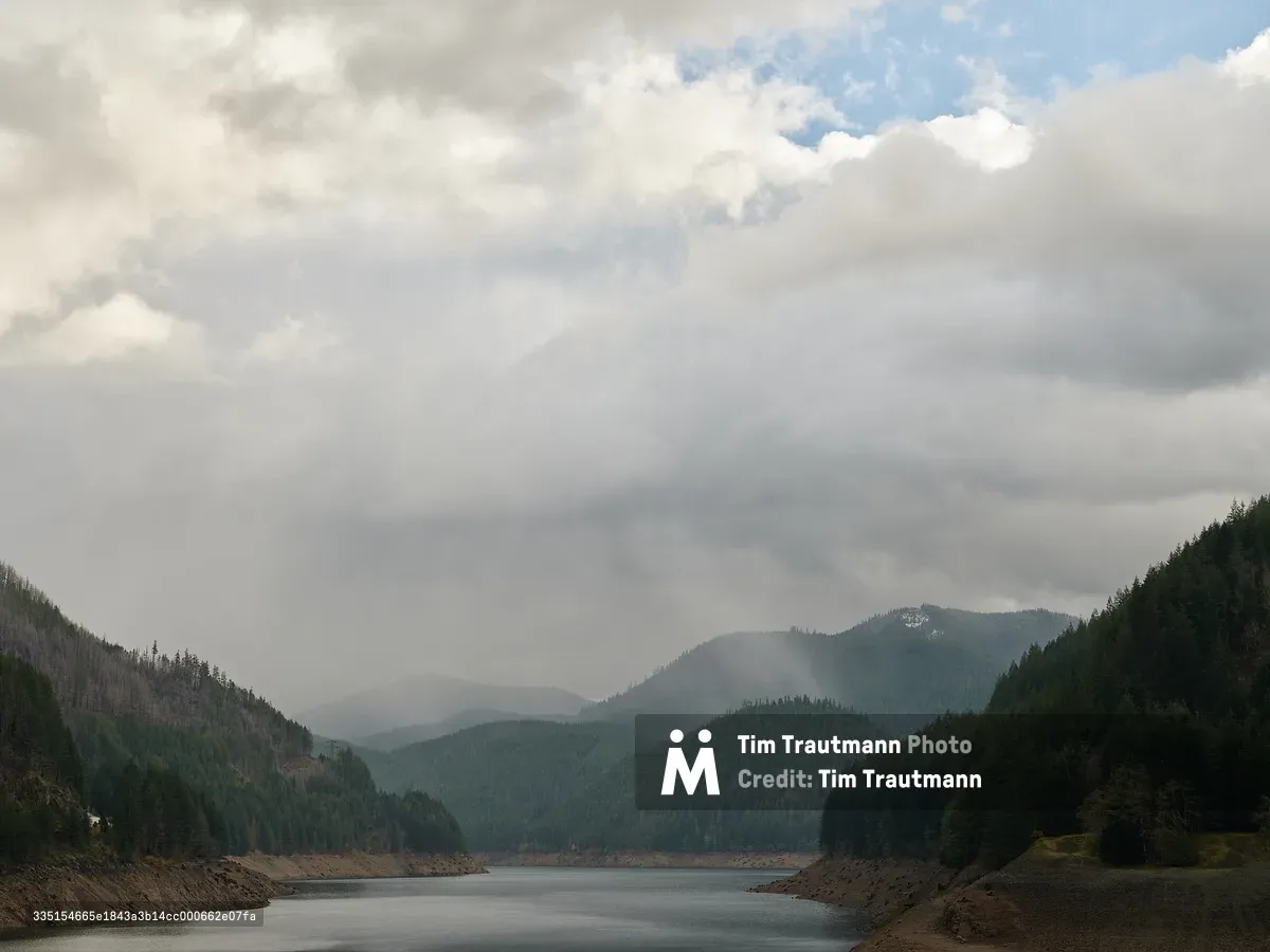 This is a moody, atmospheric landscape shot of Detroit Lake in Oregon. The reservoir stretches into the middle distance, flanked on both sides by steep, densely forested hillsides of Douglas fir and pine. The water level appears notably low, with exposed rocky and muddy shoreline visible along the banks - likely reflecting seasonal drawdown or drought conditions.  The surrounding hills show a mix of healthy green conifers and what appear to be fire-damaged or dead trees on the left slope, a reminder of the wildfires that have affected this region in recent years.  The sky dominates the upper two-thirds of the frame and is dramatic — heavy, layered storm clouds roll in from the left, with soft light breaking through on the right side, hinting at blue sky barely visible above. Mist or light rain obscures the middle mountains in the background, giving the scene a brooding, Pacific Northwest character.  The overall palette is cool and muted - slate grays, deep forest greens, and earthy browns - which gives the photo a quiet, contemplative feel. It looks like it was taken from the dam or a viewpoint above the lake, offering a clear line of sight straight up the reservoir corridor.