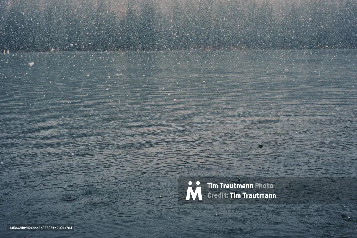 Heavy snowfall creates a curtain of white across a steel-gray lake surface in the Oregon Cascades near Sisters. The atmospheric precipitation transforms the water into a textured canvas of ripples and droplets, while the distant shoreline dissolves into a soft, monochromatic haze. The image captures the raw intensity of a mountain winter storm, with each snowflake visible as delicate points of light against the moody, overcast sky.