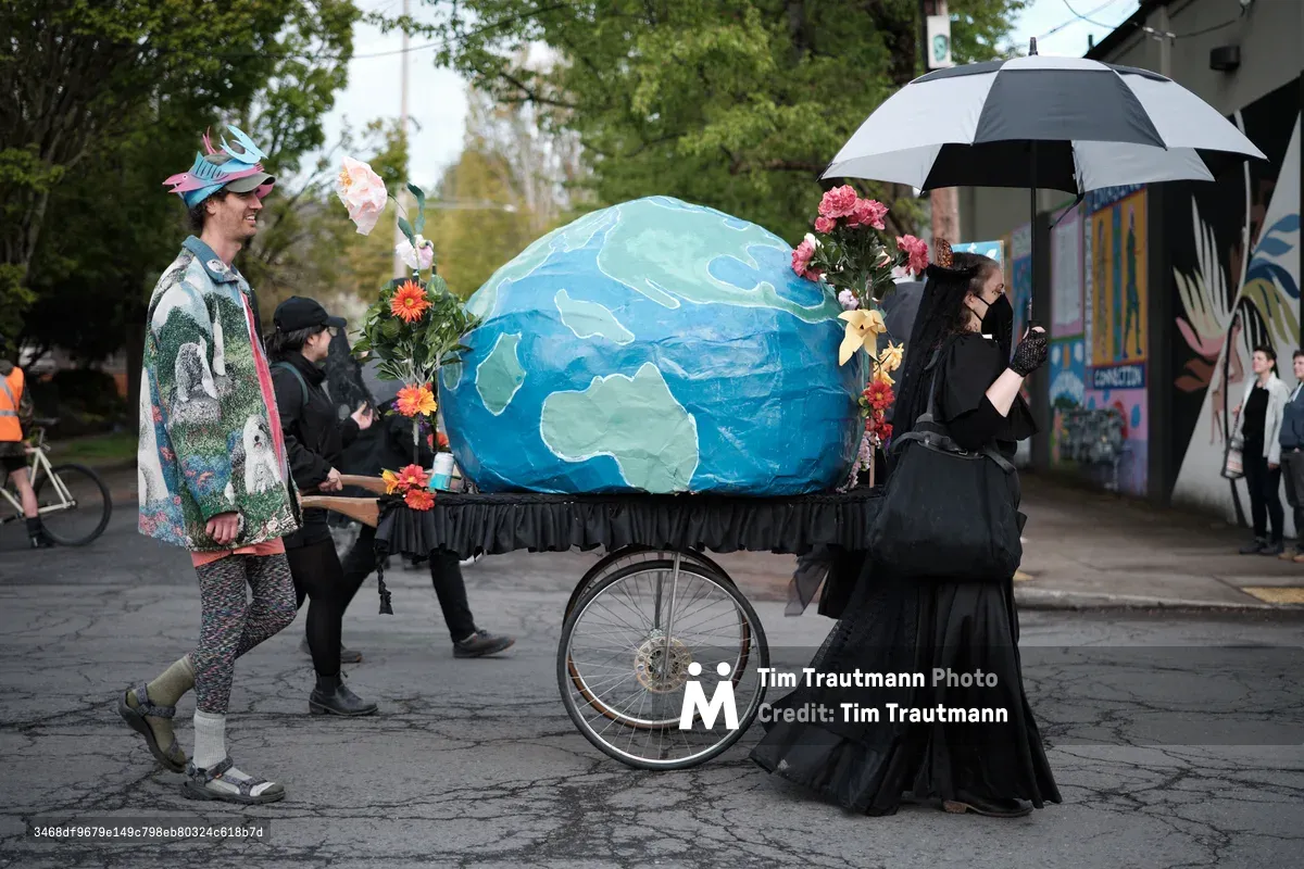 A group of environmental activists in Portland, Oregon participate in a street demonstration, pushing a wheeled cart carrying a large handmade model of Earth decorated with flowers. The protesters are dressed in colorful, eclectic clothing and one holds an umbrella, suggesting a community-organized environmental awareness event.