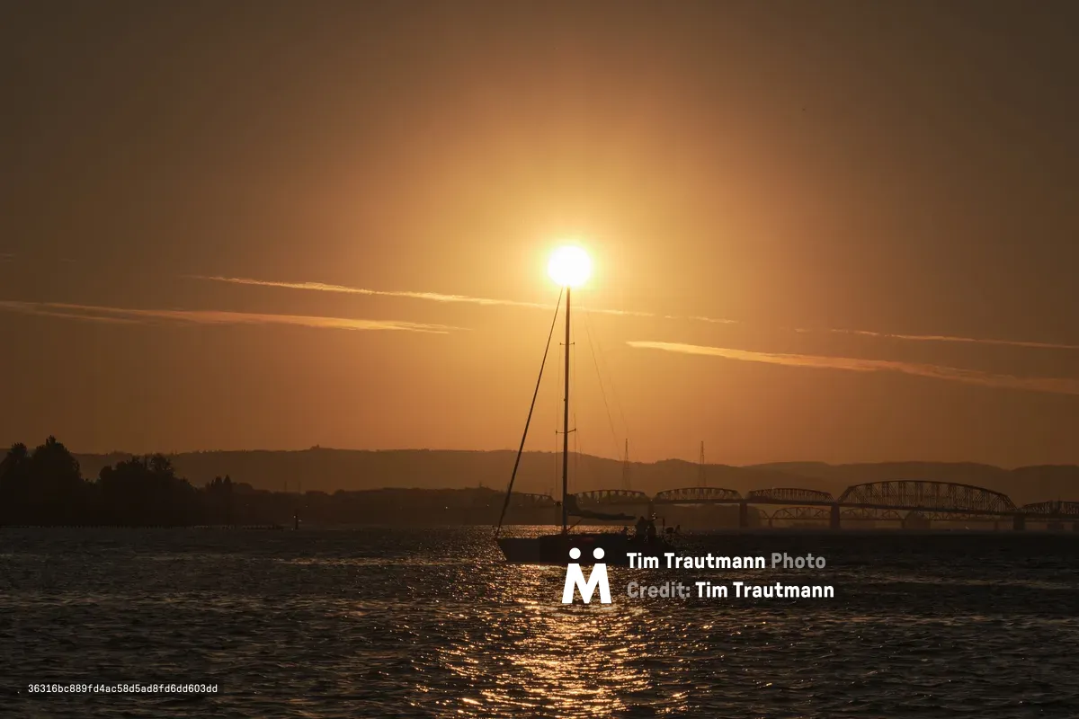 A silhouetted sailboat floats on calm water during golden hour sunset, with a large bridge visible in the background and the Columbia River reflecting warm sunlight in Portland, Oregon.