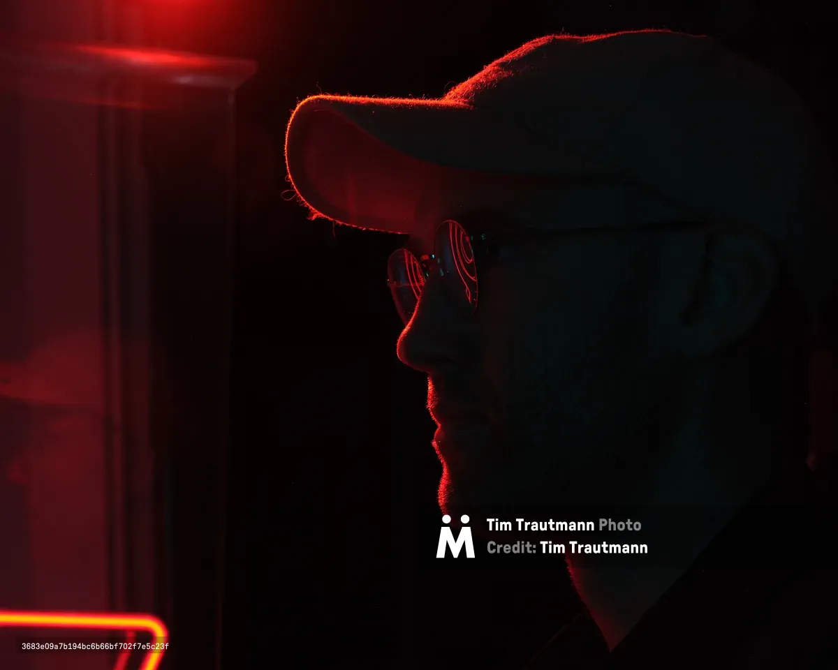 A dramatic silhouette of a person wearing a hat and sunglasses, illuminated by intense red neon lighting against a dark background. The moody lighting creates strong contrast and rim lighting effects around the subject's profile.