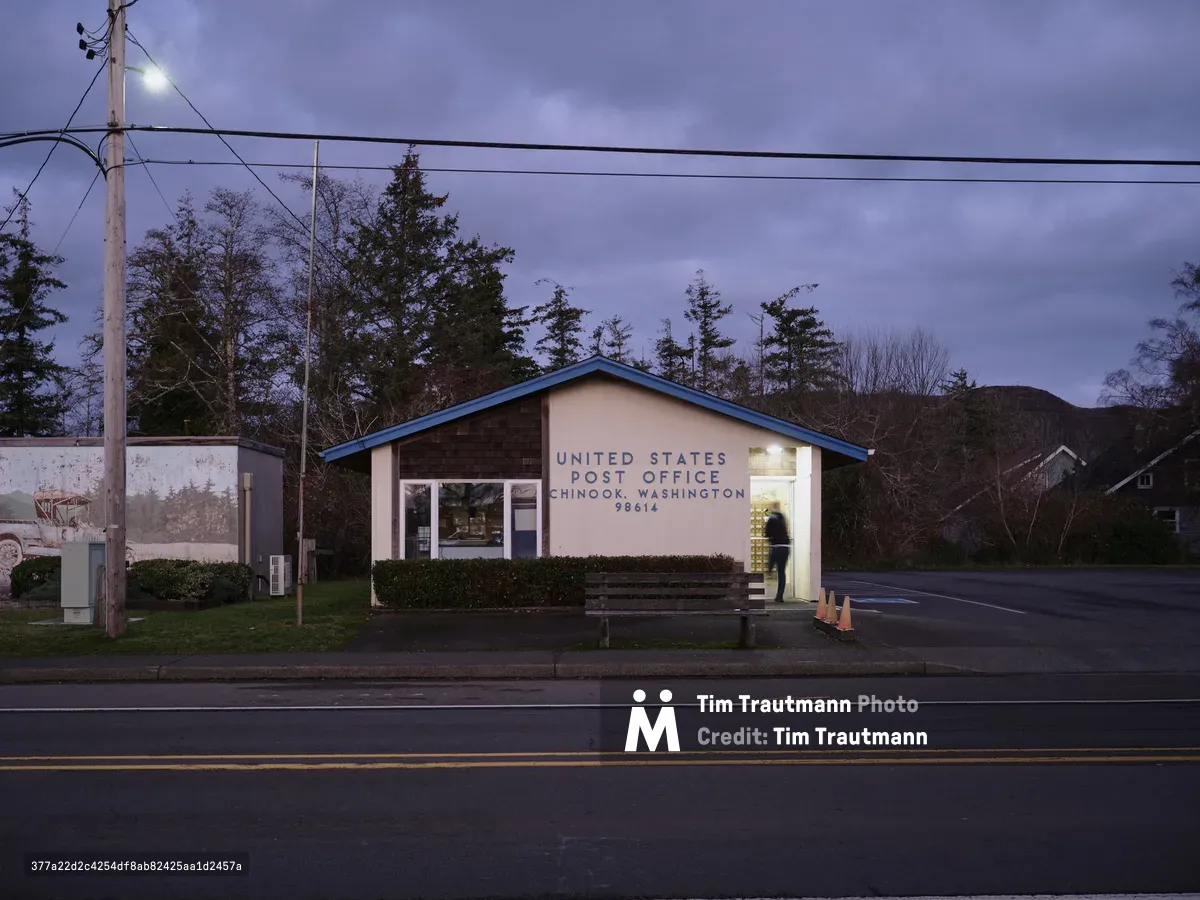 The modest United States Post Office in Chinook, Washington stands illuminated against a brooding Pacific Northwest sky, its warm interior light spilling through glass doors onto the quiet street. Power lines stretch across the frame while towering evergreens fade into the purple dusk behind the simple brick and stucco building. A solitary figure moves within the lit postal facility, creating an intimate beacon of community service in this remote coastal town where Highway 101 meets the Columbia River estuary.