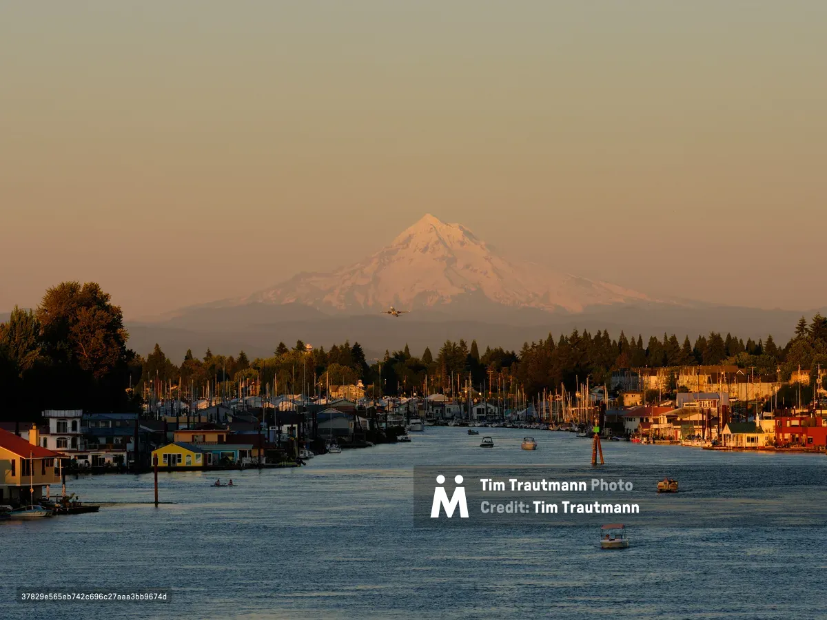 Mount Hood rises majestically through the amber-tinted atmosphere as golden hour light bathes Portland's North Harbor along the Columbia River. The snow-capped volcanic peak provides a dramatic backdrop to the bustling waterfront community, where colorful houseboats and maritime structures line the calm waters near Portland International Airport. Small aircraft can be seen ascending through the warm, hazy sky, while pleasure boats dot the serene river channel that flows between forested shores.