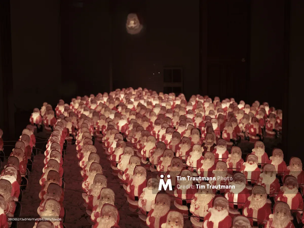 Hundreds of identical chocolate Santa figurines create a mesmerizing sea of red and white in Chris Willis's annual Portland art installation. The miniature holiday figures stretch across the darkened gallery floor in perfect rows, their faces catching warm artificial light that casts gentle shadows throughout the space. Above the sculptural army, a single moon-like orb glows mysteriously in the black void, creating an otherworldly atmosphere that transforms familiar Christmas imagery into something both whimsical and haunting.