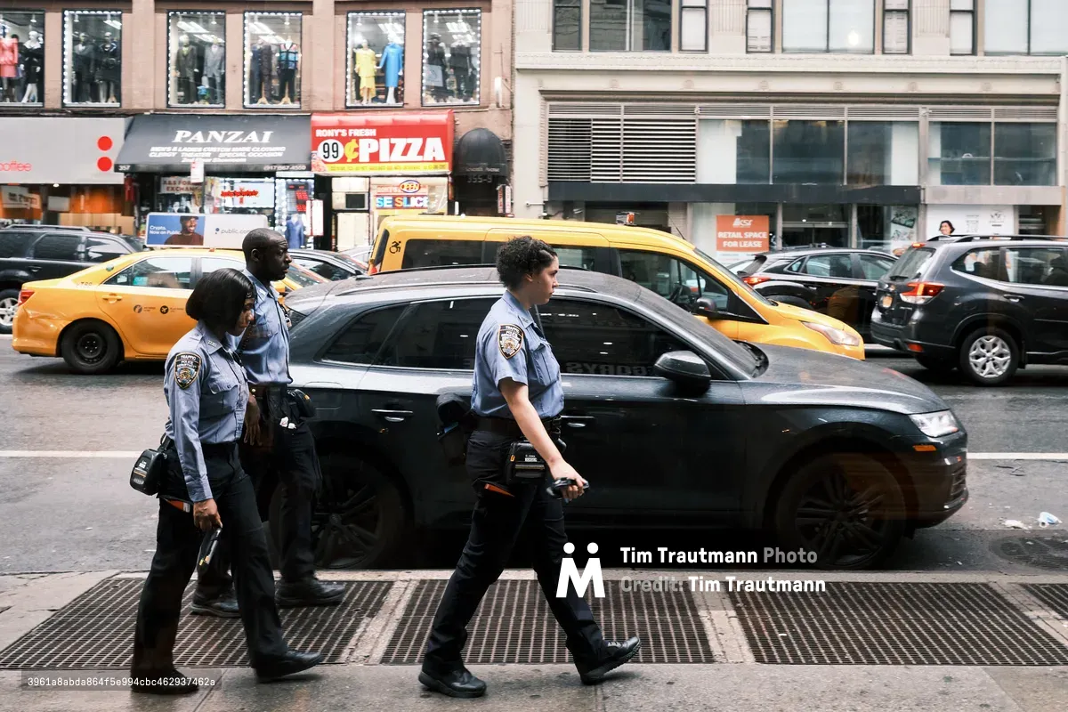 Three uniformed police officers stride purposefully along 7th Avenue in Manhattan's Chelsea District, their light blue shirts and dark equipment belts catching the filtered daylight. Behind them, yellow taxi cabs and a sleek black sedan are locked in typical New York traffic, while storefronts including a pizza joint and clothing displays create the quintessential urban backdrop. The scene captures the rhythm of city law enforcement moving through the congested arteries of midtown Manhattan, where pedestrian and vehicular worlds intersect in perpetual motion.
