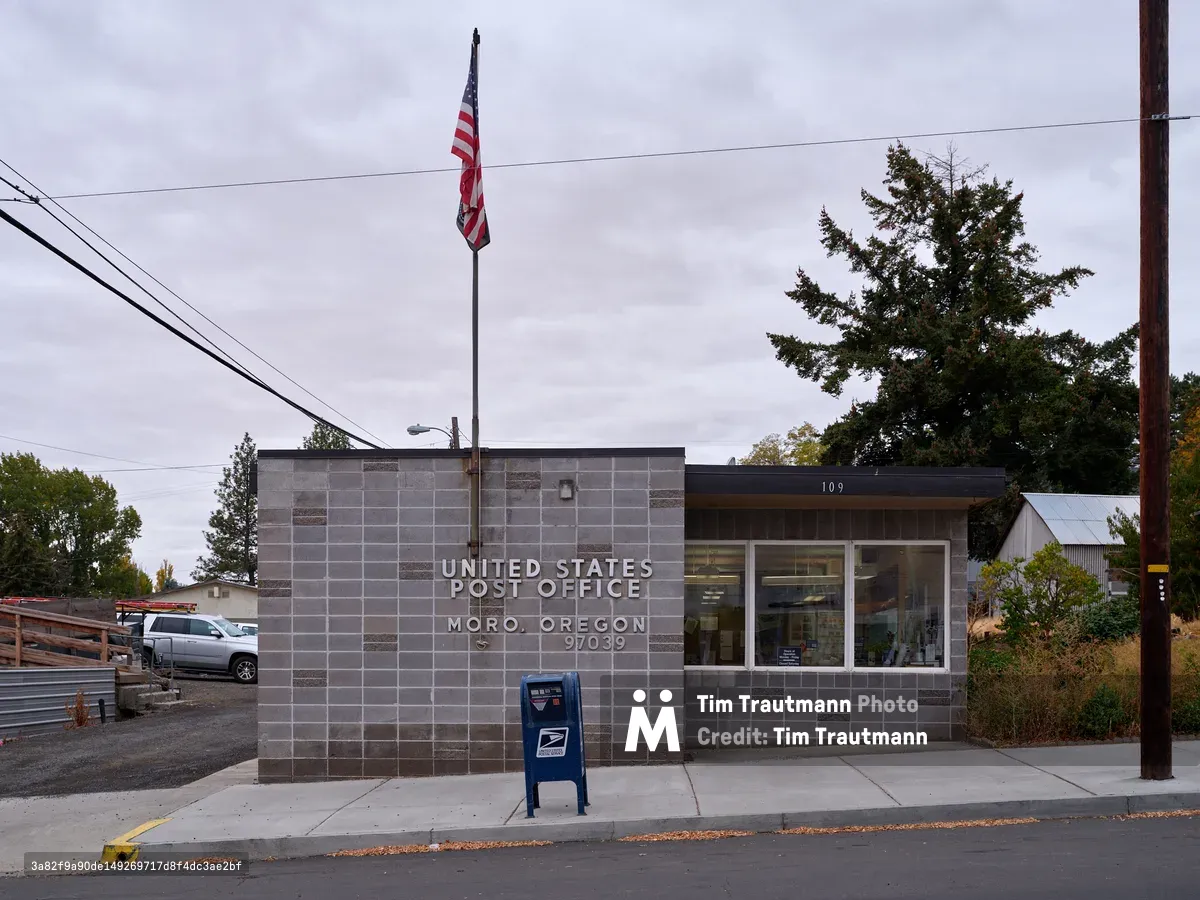 A contemporary United States Post Office building stands quietly along 1st Street in Moro, Oregon, its gray concrete block construction reflecting the utilitarian architecture of rural America. The American flag waves solemnly from its pole under a heavy overcast sky, while power lines stretch across the frame, emphasizing the small-town infrastructure. A classic blue USPS collection box stands sentinel on the sidewalk, and the building's clean modernist lines contrast with the mature evergreen trees that frame the scene in this Sherman County community.