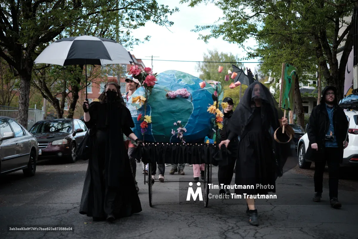 A group of protesters dressed in black robes march down a tree-lined street in Portland's Central Eastside district, carrying a large blue Earth globe decorated with flowers on a wheeled cart. The solemn procession appears to be an environmental demonstration or funeral-themed climate protest, with participants holding umbrellas and wearing dark clothing while walking among parked cars on Southeast 14th Avenue.