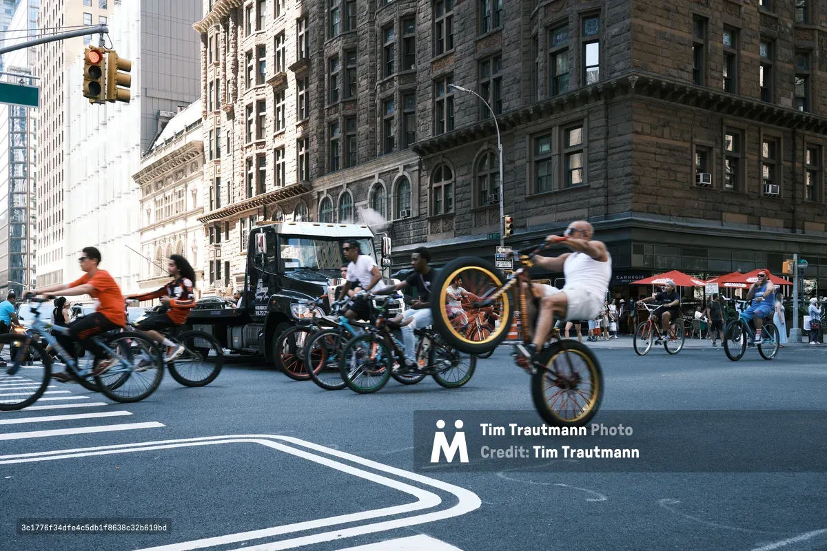 A dynamic stream of cyclists floods the asphalt canyon of 7th Avenue in Midtown Manhattan, their motion-blurred forms creating a river of urban energy against the stoic backdrop of historic brownstone facades and towering glass. The late afternoon light catches the warm tones of the pre-war architecture while modern skyscrapers pierce the frame, creating a layered narrative of New York's architectural evolution. A vintage-style truck anchors the composition as pedestrians and outdoor diners bear witness to this spontaneous parade of two-wheeled commuters.