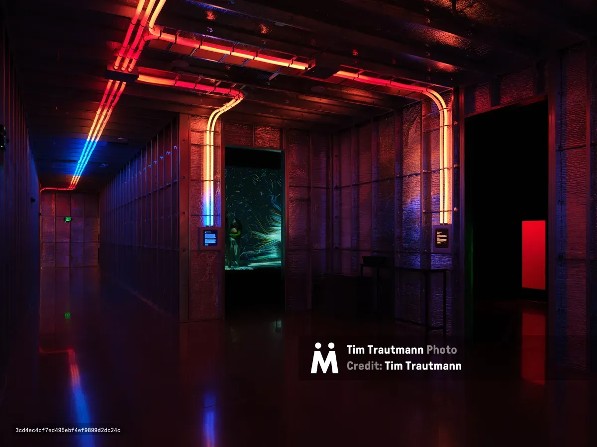 A dramatic interior hallway at Hopscotch venue in Portland featuring exposed brick walls, industrial ceiling fixtures, and vibrant neon lighting in red, blue, and green creating atmospheric reflections on the polished floor. The space showcases modern industrial design with color-changing LED tubes and digital displays along the corridor.