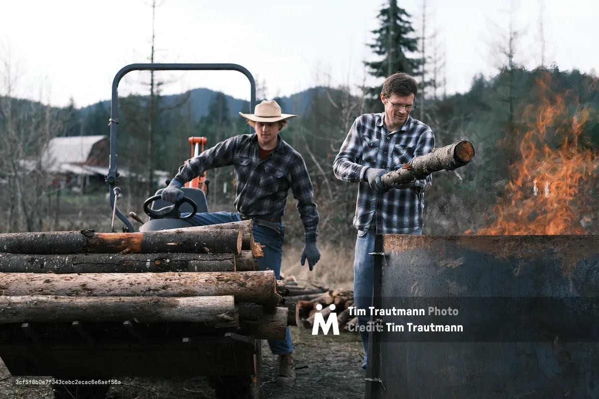 Two farmers in matching plaid flannel shirts and work gloves make biochar on a rural property. The man on the right feeds a log into a large metal burn barrel with open flames, while the younger man on the left, wearing a straw cowboy hat, tends to a pile of logs on a trailer. A chainsaw rests on the trailer nearby. A barn, bare-limbed trees, and forested hills are visible in the background under an overcast sky.