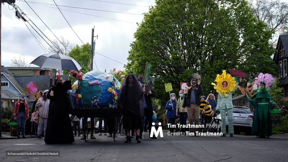 A community protest march on a residential Portland street featuring participants in costumes including someone dressed as the Grim Reaper, people carrying a large inflatable Earth globe decorated with flowers, and others wearing flower costumes representing environmental themes. The diverse group of protesters walks down Southeast Salmon Street in the Buckman neighborhood under an overcast sky.
