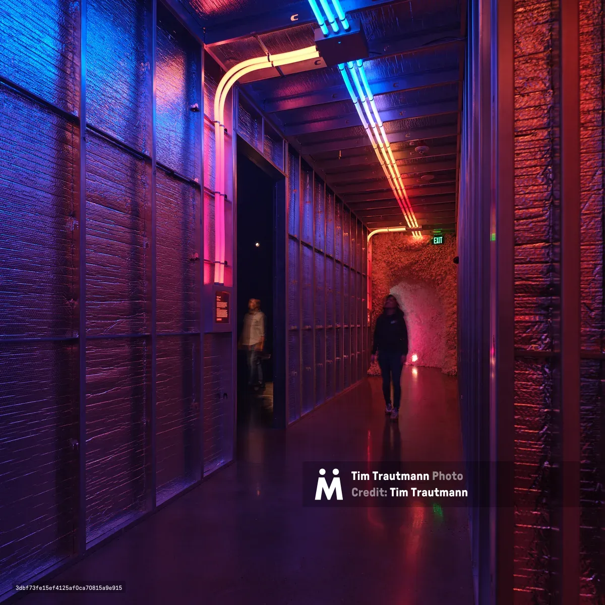 A dramatically lit hallway at Hopscotch entertainment venue in Portland, Oregon, featuring exposed brick walls illuminated by vibrant blue and red neon lighting. Two silhouetted figures walk through the atmospheric corridor with its industrial ceiling and polished concrete floors.