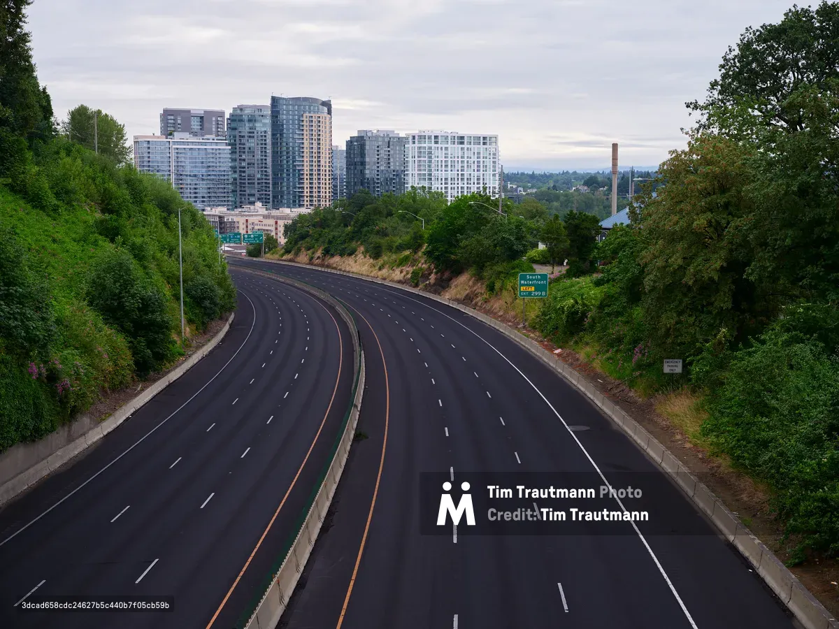 A wide, empty multi-lane highway curves through lush green hills toward the modern high-rise buildings of downtown Portland, Oregon under an overcast sky. The scene captures the contrast between urban development and natural landscape typical of the Pacific Northwest.
