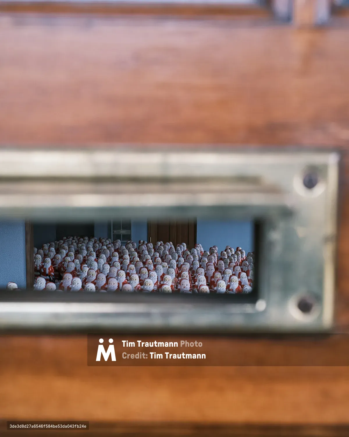 Through the narrow aperture of a gallery mailslot, hundreds of identical Santa figurines crowd together in ghostly formation, their red-suited forms creating a sea of holiday repetition. The voyeuristic perspective frames this uncanny army of Christmas icons within the confines of Portland's Consignment Gallery, where artist Chris Willis's annual installation transforms the familiar into the surreal. Warm wood tones of the gallery exterior contrast sharply with the cool interior lighting that illuminates this manufactured multitude, creating an intimate yet unsettling glimpse into mass-produced festivity.