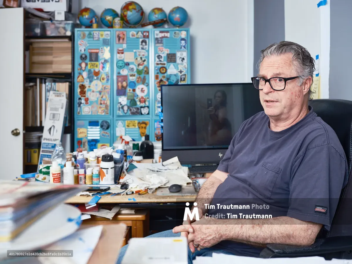 A middle-aged man with glasses sits in his cluttered creative studio workspace, surrounded by art supplies, globes, and colorful displays on the walls. The space shows an active artist's environment with paints, papers, and various creative materials scattered across his desk.