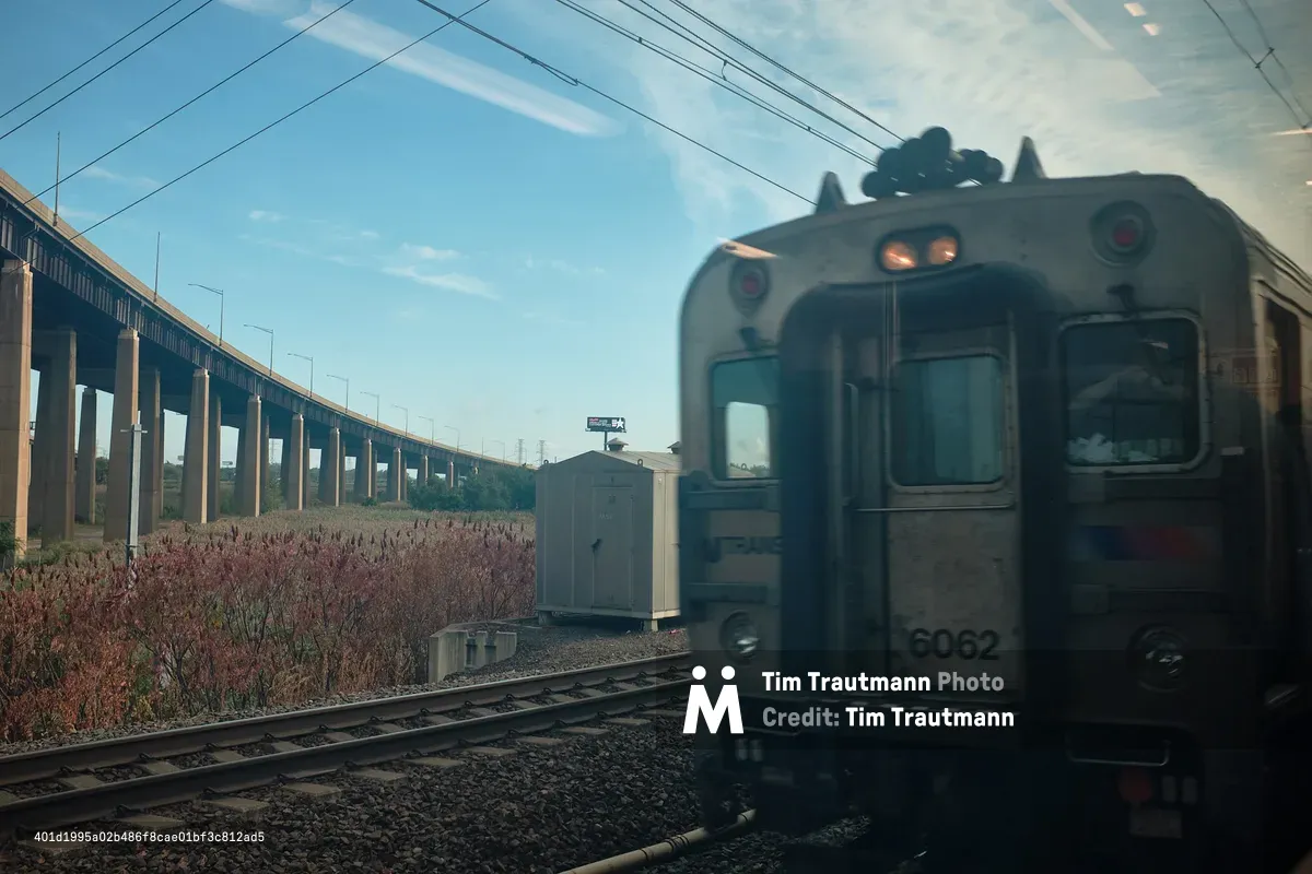 A close-up view of an NJ Transit commuter train car numbered 6062 passing on an adjacent track, photographed through a window near Kearny, New Jersey. A conductor is visible leaning against the glass inside the passing train. Wild reddish-pink vegetation lines the rail corridor in the foreground. A large elevated Interstate 95 viaduct stretches across the background on concrete columns under a blue sky with wispy clouds. A small utility shed and a distant billboard are visible between the train and the highway overpass.