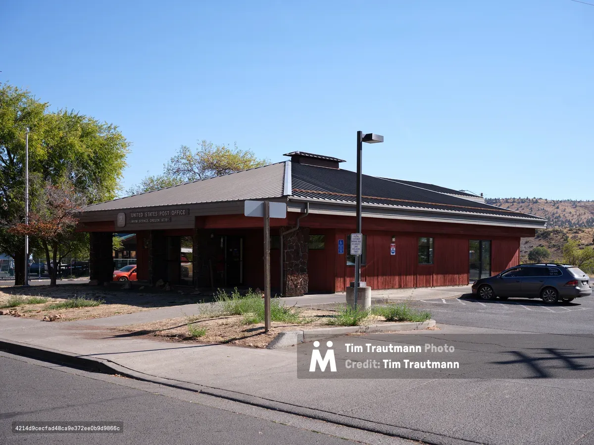 A weathered brick United States Post Office stands as a community anchor in Warm Springs, Oregon, its earth-toned facade harmonizing with the high desert landscape. The low-slung building features a distinctive overhanging roof and covered entrance, casting deep shadows across the front walkway under the brilliant blue sky. Scattered vehicles and sparse vegetation speak to the quiet rhythm of small-town life, while distant hills create a backdrop of muted autumn colors.