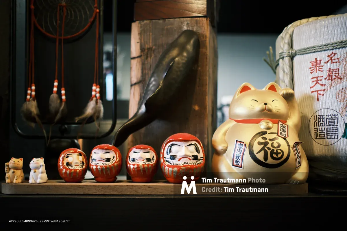 A curated arrangement of Japanese good luck talismans creates an intimate shrine within Portland's Sushi Mazi restaurant. Four vermillion daruma dolls sit in perfect progression alongside a golden maneki-neko cat, while traditional rope-bound sake containers and ceramic vessels frame the composition in warm, ambient lighting. The weathered wooden shelf and shadowy depths behind speak to authenticity and reverence, transforming everyday restaurant décor into a meditation on prosperity and protection.