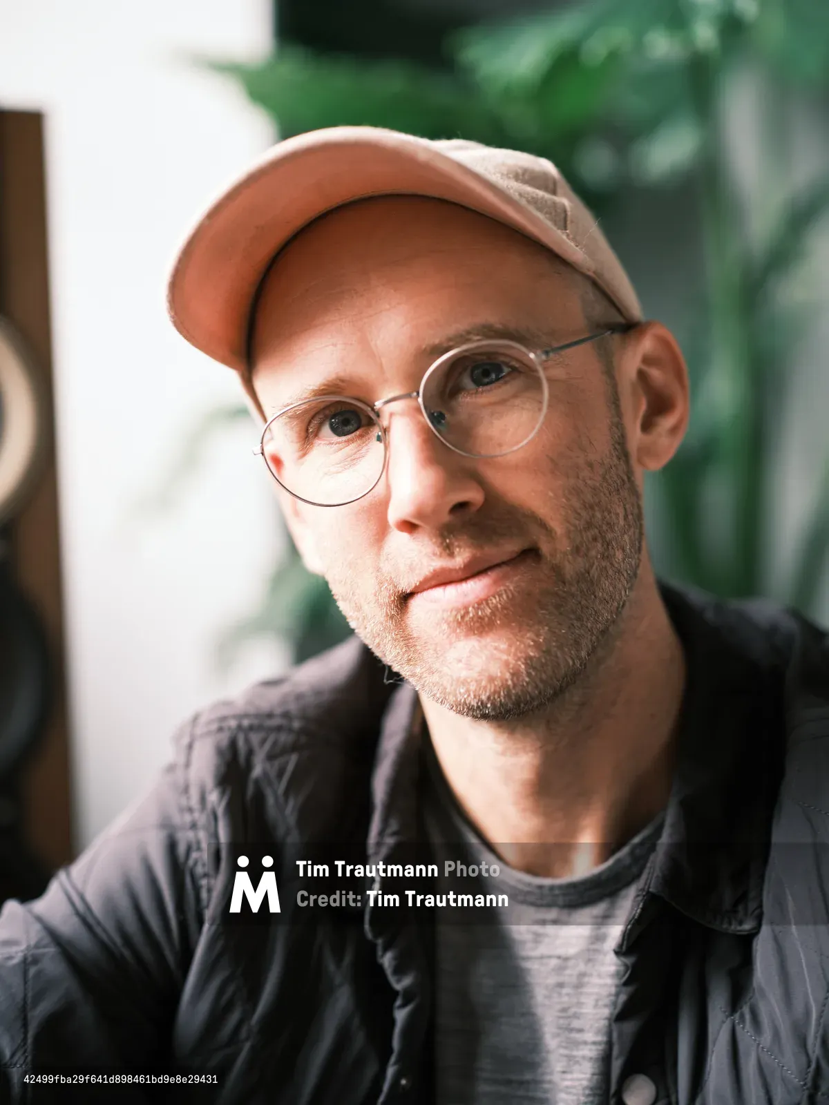 A warm portrait of a middle-aged man wearing round glasses, a tan baseball cap, and dark jacket, smiling gently at the camera with soft natural lighting and greenery in the background.
