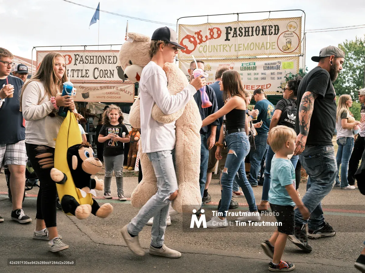A bustling crowd of fairgoers walks through the Clark County Fair in Vancouver, Washington. In the center of the frame, a teenage boy in a white sweatshirt and black cap carries an enormous stuffed teddy bear he won at a carnival game. To his left, a girl carries a large stuffed monkey in a banana costume. Families, children, and adults fill the midway, with food vendor signs for Old Fashioned Country Lemonade and Old Fashioned Kettle Corn visible in the background. Several people carry colorful fair drinks.