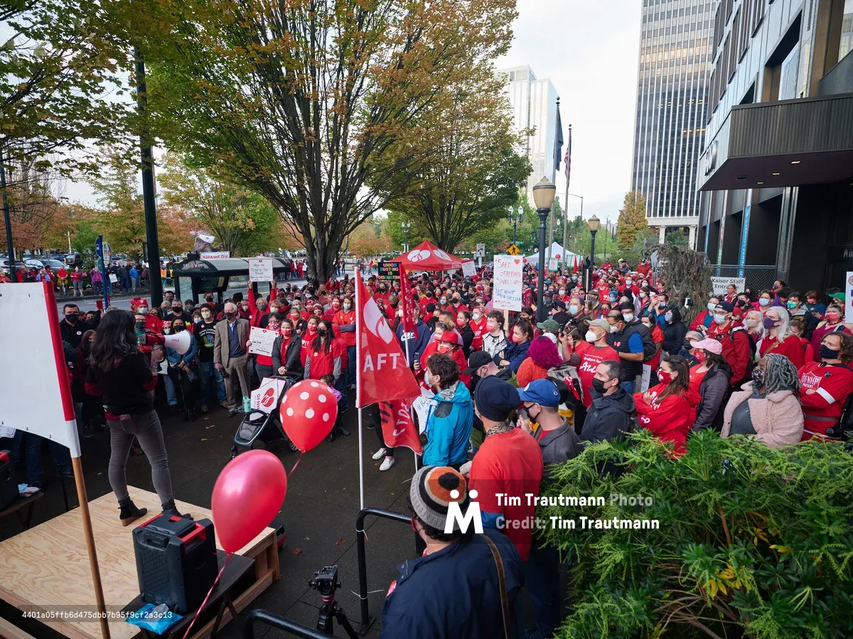 Hundreds of nurses and healthcare workers in vibrant red union attire congregate outside Kaiser Permanente's towering glass headquarters in Portland's Lloyd District, their determined faces reflecting the gravity of impending labor action. The autumn foliage of street trees creates a golden canopy above the crowd, while red AFT banners and protest signs punctuate the sea of solidarity. A makeshift speaking platform commands attention from the diverse assembly, capturing the pivotal moment before a significant healthcare strike unfolds against the urban backdrop of downtown Portland.