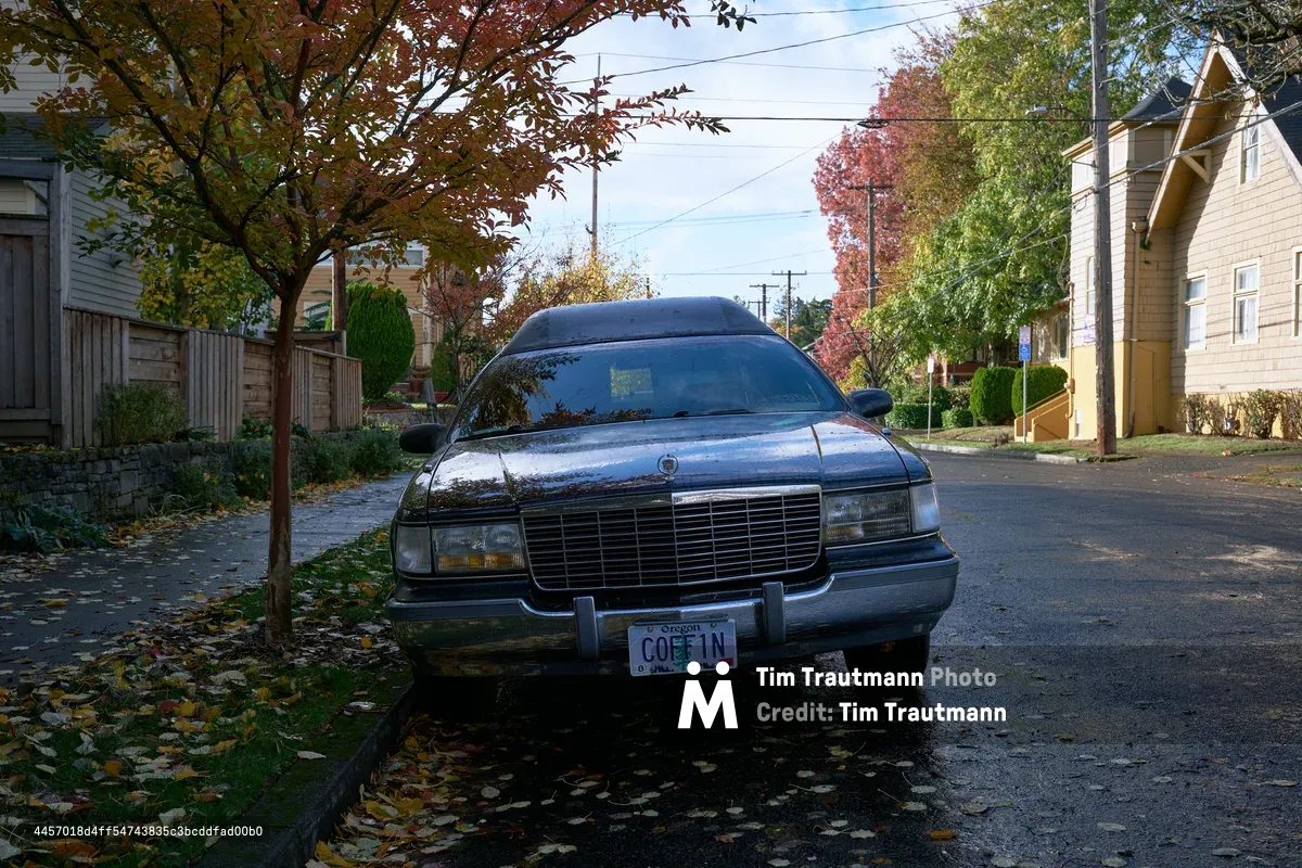 A black vintage hearse with Oregon license plate 'COFFIN' is parked on a wet asphalt street in a residential Portland neighborhood during autumn. The scene features colorful fall foliage, wooden fences, and characteristic Pacific Northwest housing architecture.