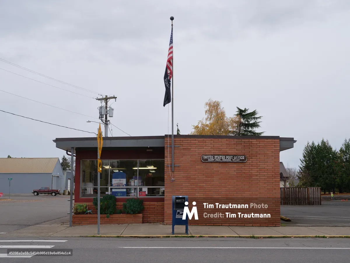 A modest brick United States Post Office stands sentinel in Saint Paul, Oregon, its mid-century modern architecture crowned by the American flag and POW-MIA banner fluttering against pewter-gray clouds. The building's warm red brick facade contrasts with the somber autumn sky, while a lone blue mailbox guards the sidewalk and utility lines sketch geometric patterns overhead. The scene captures the quiet dignity of rural American infrastructure, where community services endure beneath the Pacific Northwest's characteristic overcast atmosphere.