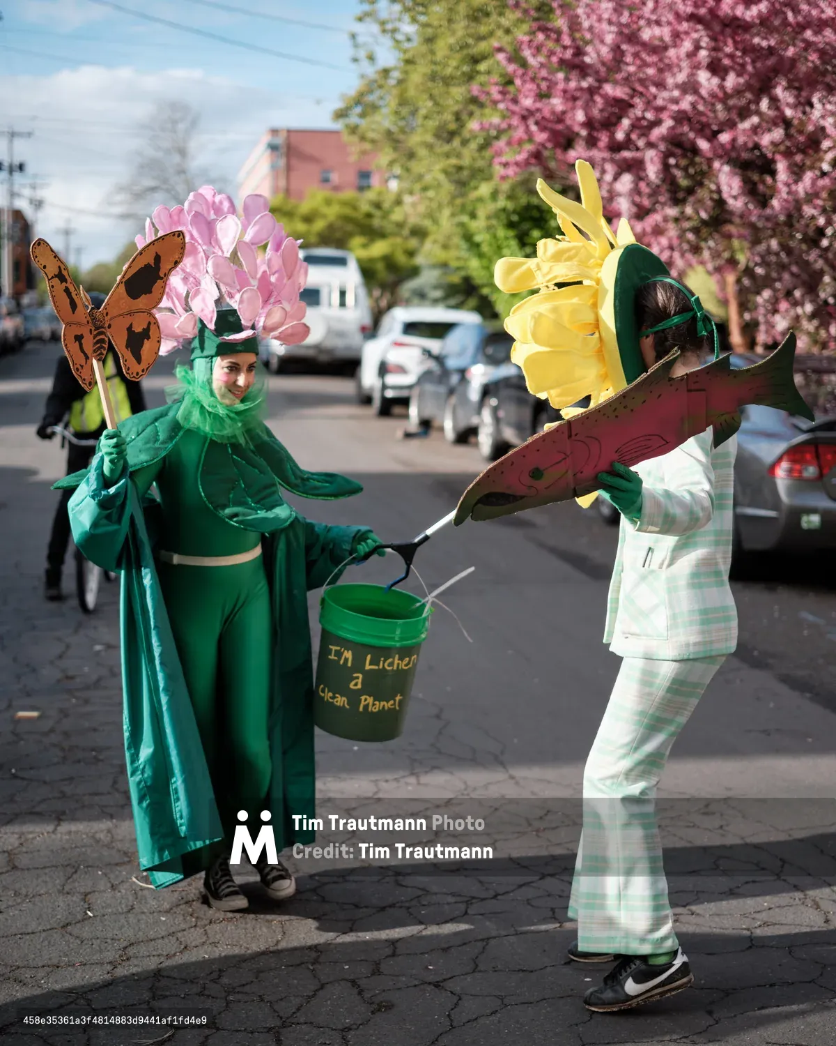 Two costumed street performers in Portland, Oregon engage in an environmental awareness demonstration, with one person dressed as a flower holding a bucket labeled 'I'm Lichen a Clean Planet' and another wearing nature-themed props including butterfly and fish cutouts.