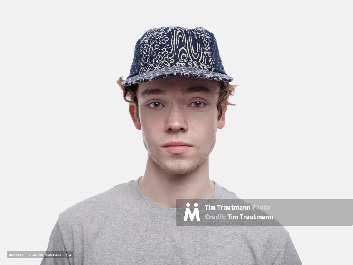 A young man with tousled blonde hair gazes directly at the camera with an earnest expression, wearing a navy blue paisley-patterned baseball cap and heather gray t-shirt. The portrait is captured against a clean white backdrop with even, diffused lighting that creates a soft, contemporary mood. His relaxed posture and steady gaze suggest casual confidence, while the intricate paisley design on his cap adds visual texture and cultural reference to the otherwise minimalist composition.